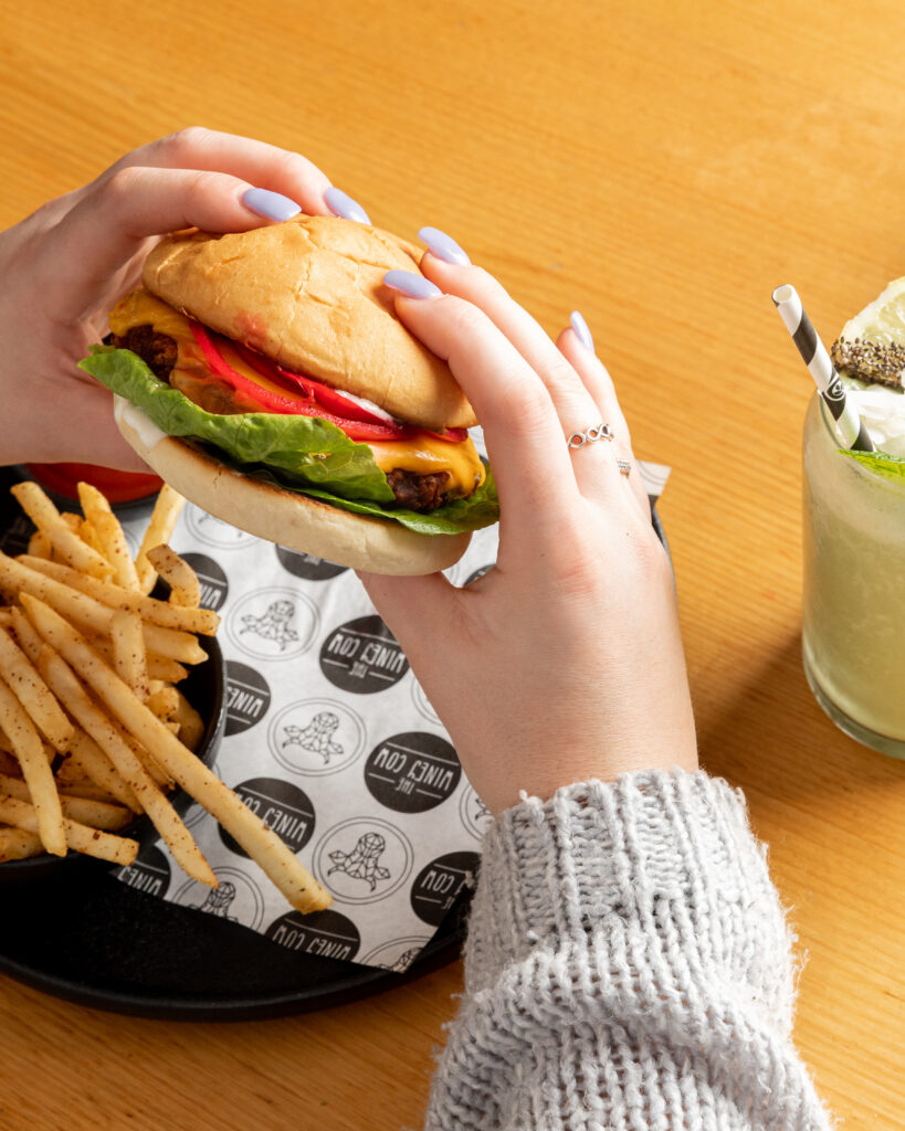 A photo of two hand holding a colourful veggie burger, with a delicious and bright green smoothie sitting on the table.