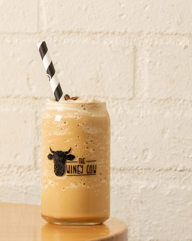A photo of a golden iced coffee in a branded Winey Cow glass with a striped straw, sitting on a wooden table with a white brick background.
