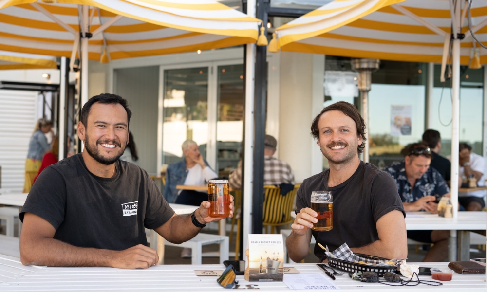 A photo of two men sitting outside under yellow umbrellas with beers in their hands.