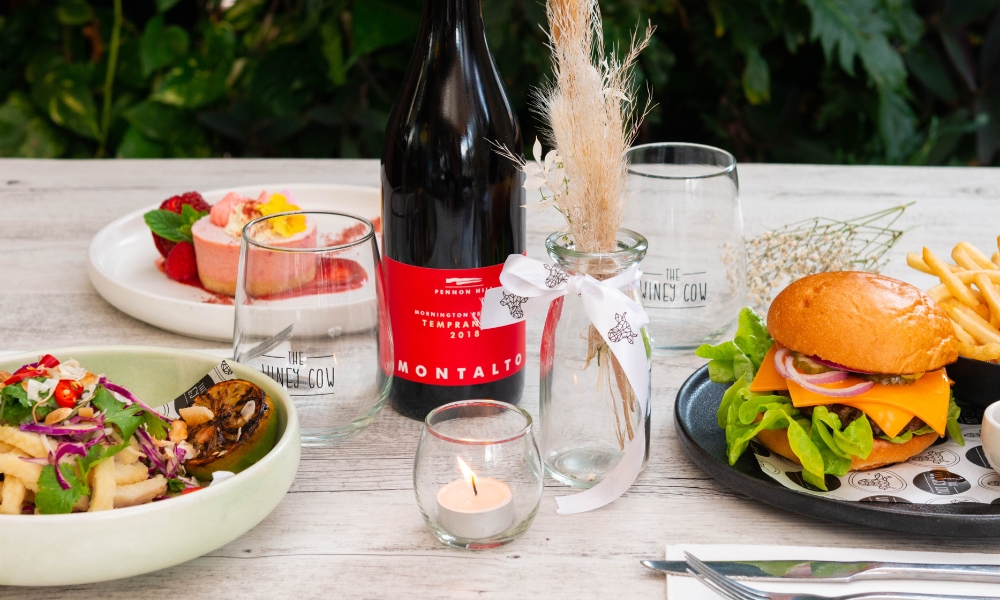 A beautiful table setting with a bottle of wine, two meals, a dessert and a candle.