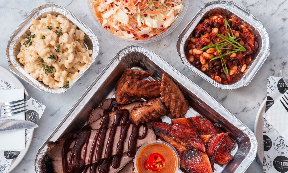 A bird's eye view photo of a catering pack designed for families. The dish is assorted smoked meats with sides of beans, slaw and mac & cheese.