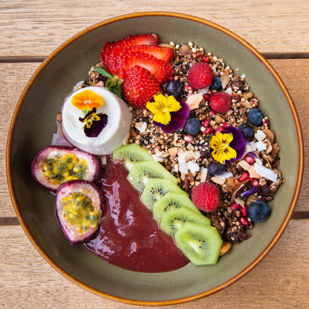 A close up photo of a colourful granola topped with kiwi fruit, passion fruit berries and a side of panna cotta.