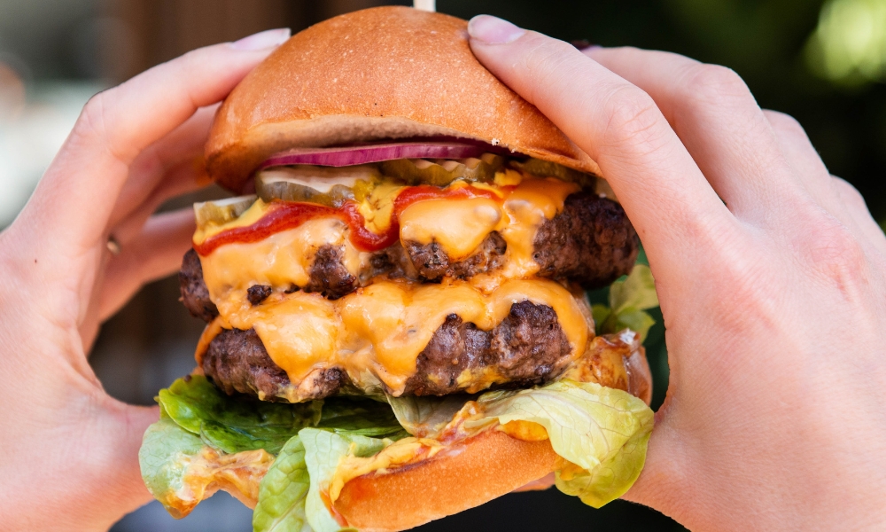 A photo of two hands holding a burger with two beef patties, dripping cheese and lettuce in between two buns.