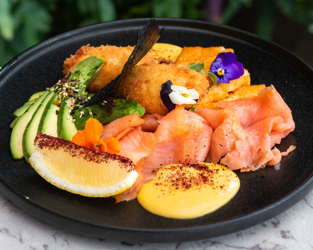 A photo of a breakfast dish with panko-crumbed eggs, smoked salmon, avocado and hollandaise on a black plate.