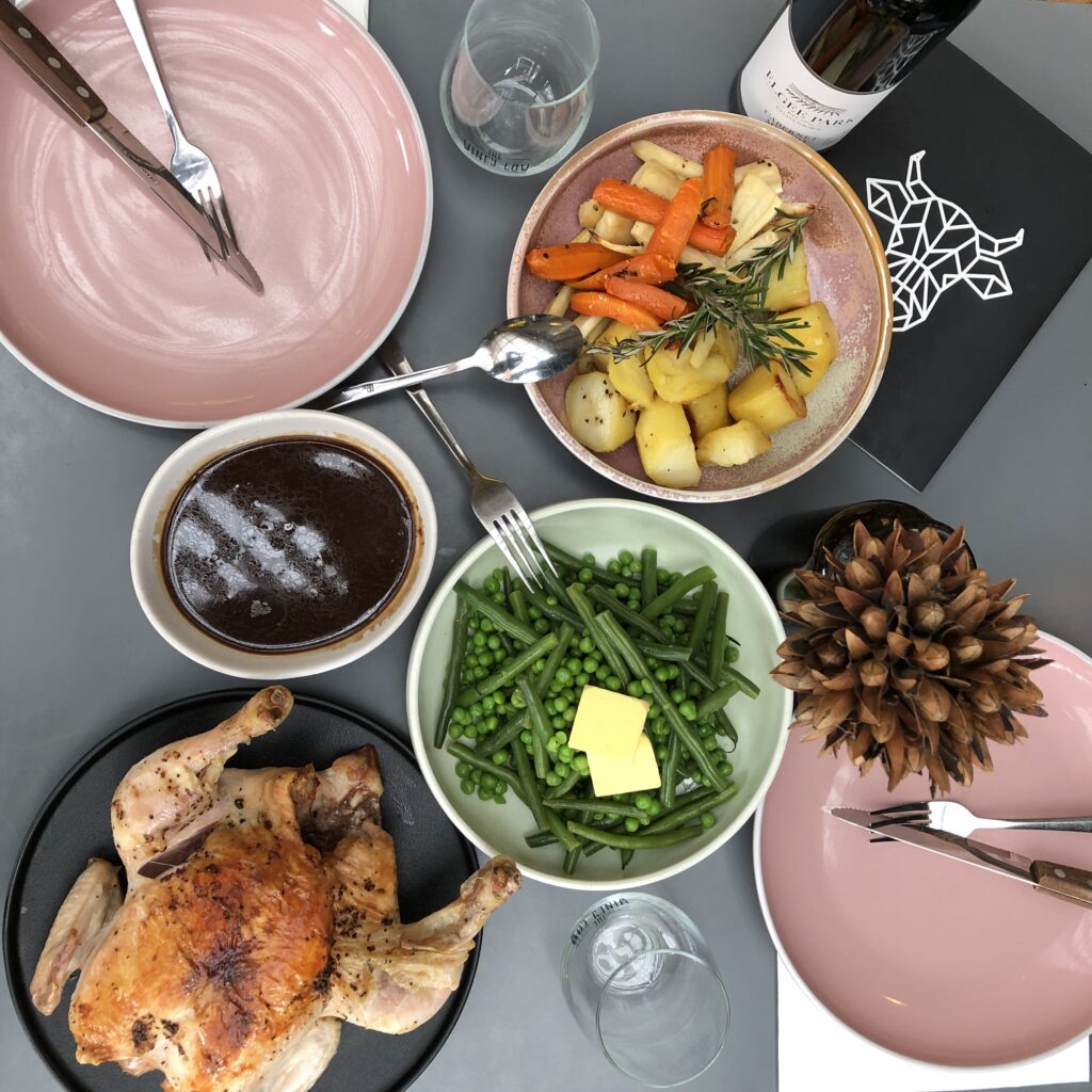 A table set with wine glasses, a bottle of red wine, and a selection of meals including a roast chicken, peas and potatoes.