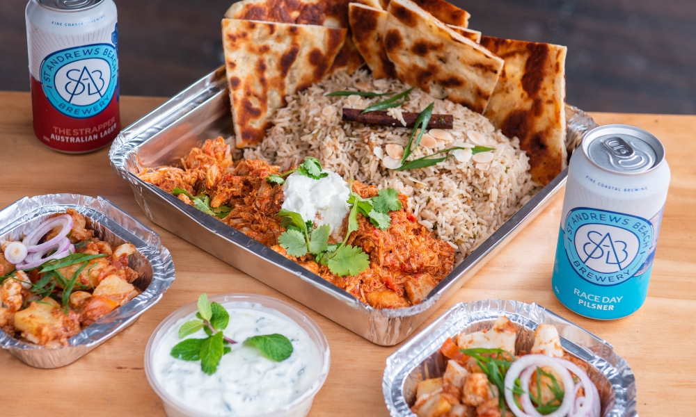 A photo of a bright Indian curry with rice served in a takeaway catering container. The dish is accompanied by some side dishes and two beers from a local brewery.