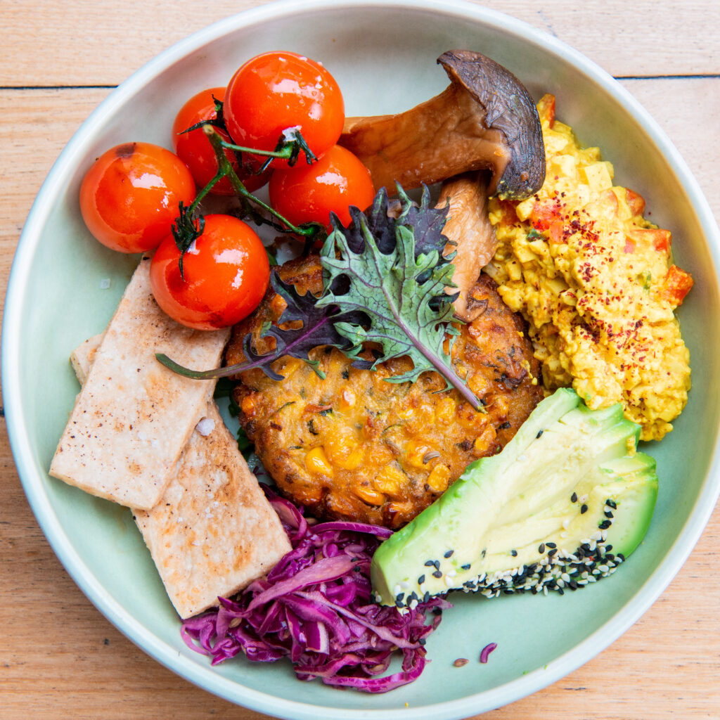 A birds-eye photo of a bowl filled with vegetarian food. It includes, tomatoes, scrambled tofu, avocado, sauerkraut and a corn & zucchini fritter.