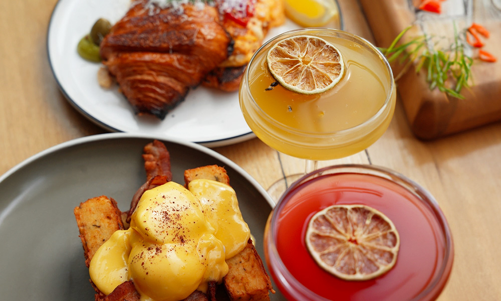 A top view perspective of a yellow and a red mimosa next to two delicious breakfast meals on a wooden table.