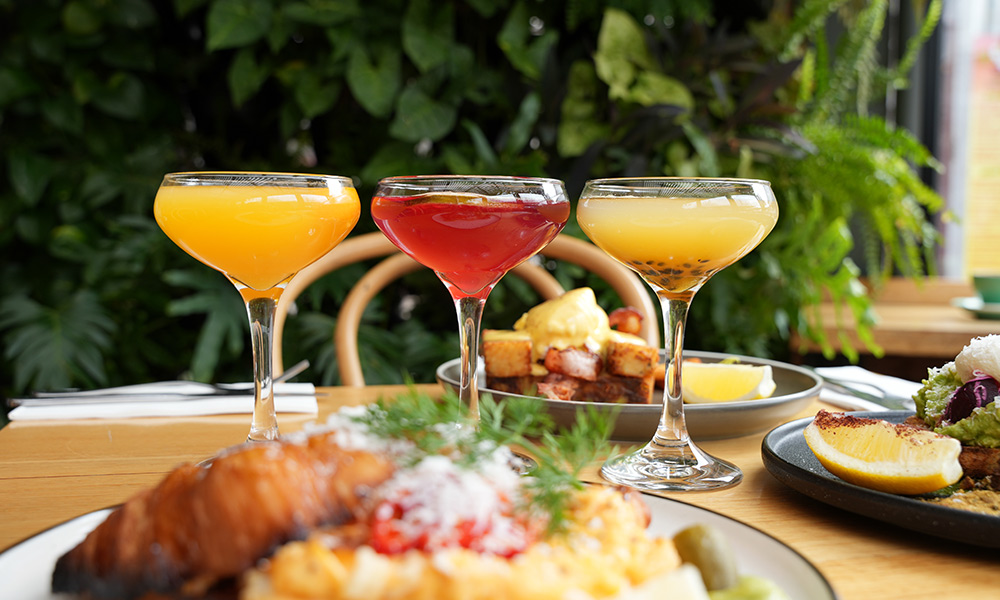 Three colorful mimosas on a wooden table next to 3 breakfast meals with greenery in the background.