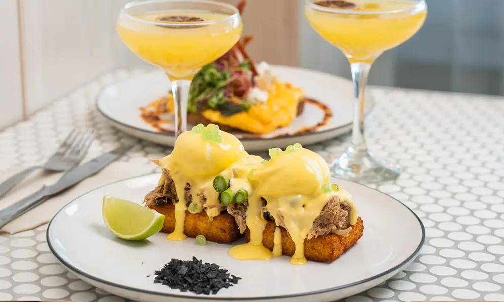 Plate of a delicious brunch stack with poached eggs, shredded meat with hollandaise dropping off the side on top of a circle tile table with 2 mimosas in the background.