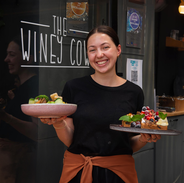 A smiling staff member of The Winey Cow holding two plates of beautifully presented dishes in front of the restaurant's sign.