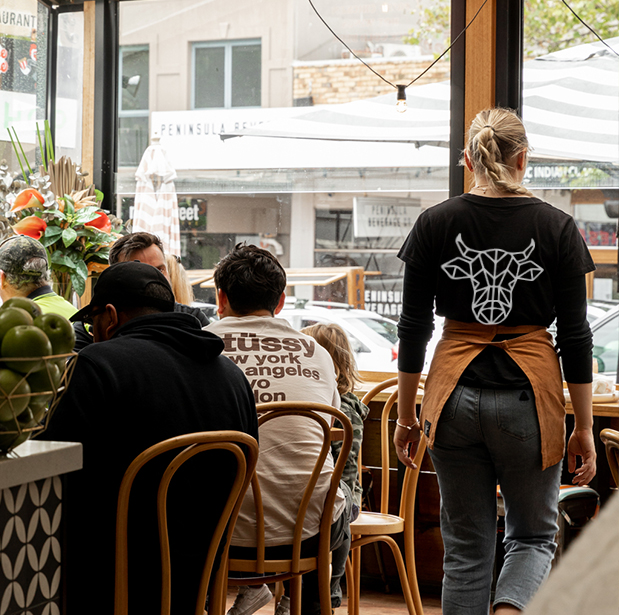 Waitress with The Winey Cow logo on her shirt walking towards customers seated inside the restaurant, with a view of the busy street outside.