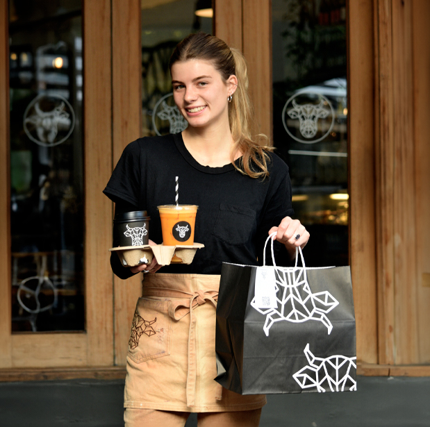 Smiling waitress holding a tray with coffee and juice alongside a takeout bag with The Winey Cow logo, standing in front of the restaurant's entrance.