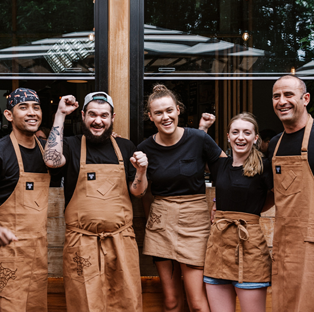 Smiling team members of The Winey Cow standing together in front of the restaurant, celebrating with raised fists and wearing brown aprons.