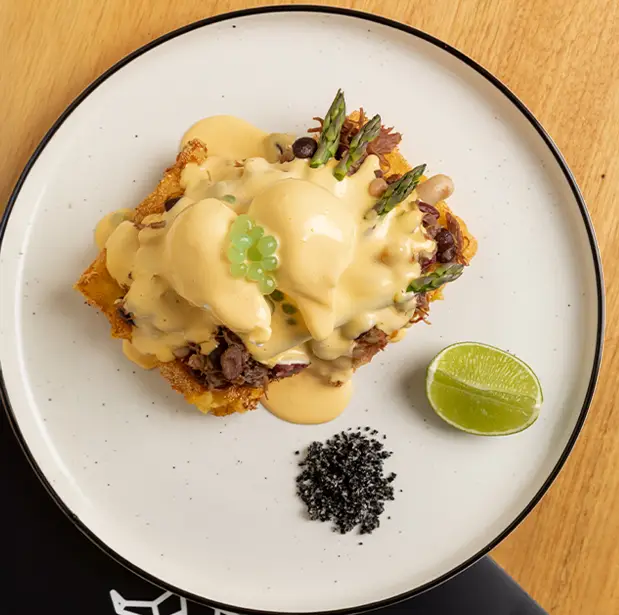 A gourmet brunch dish from The Winey Cow, showcasing poached eggs with hollandaise sauce, crispy toast, asparagus spears, and a side of lime and black salt, artfully arranged on a white plate with a black rim.