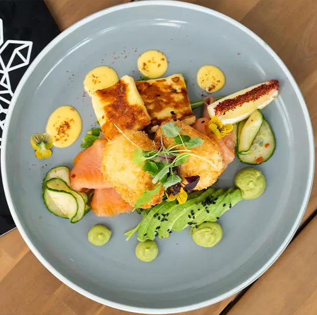 A gourmet brunch plate from The Winey Cow, featuring fried halloumi, smoked salmon, poached eggs, avocado, cucumber slices, lemon, and decorative edible flowers, arranged artistically on a grey plate.