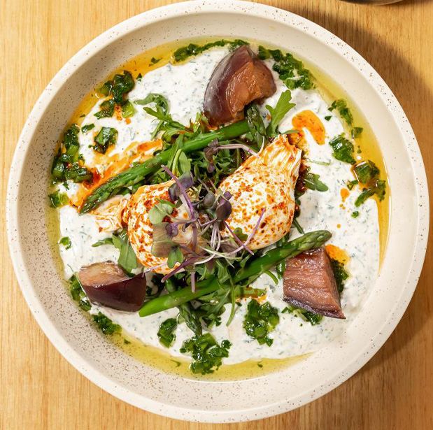 A beautifully plated dish from The Winey Cow, featuring poached eggs on a bed of fresh asparagus, greens, and a creamy herb sauce, garnished with microgreens and served in a speckled white bowl on a wooden table.