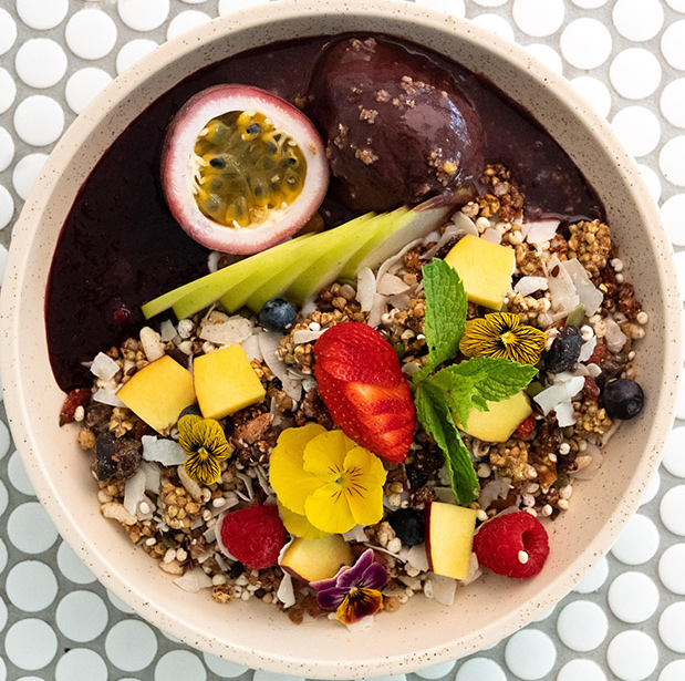 A vibrant acai bowl from The Winey Cow, topped with fresh fruit including passion fruit, mango, strawberries, and blueberries, along with granola, coconut flakes, mint leaves, and edible flowers, served in a white bowl on a patterned table.