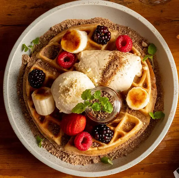 A beautifully plated waffle dish from The Winey Cow, topped with fresh berries, banana slices, two scoops of ice cream, a dollop of whipped cream, and garnished with mint leaves, served on a white plate with a light dusting of crumbled toppings.