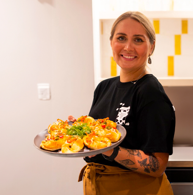 A young woman with a bright smile, dressed in a branded uniform and tan apron, serving a beautifully arranged tray of appetizers.