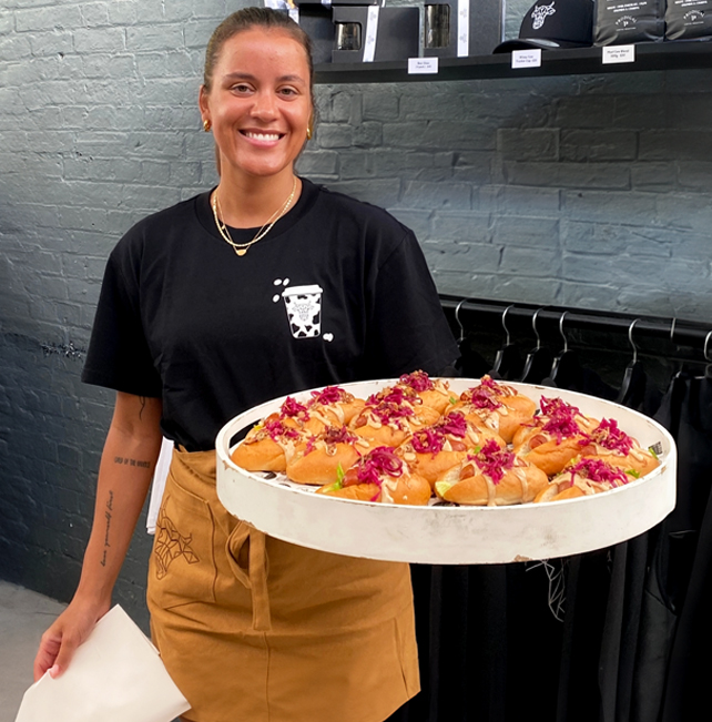 A young woman with a bright smile, dressed in a branded uniform and tan apron, serving a beautifully arranged tray of mini hotdogs to guest at a catered event.