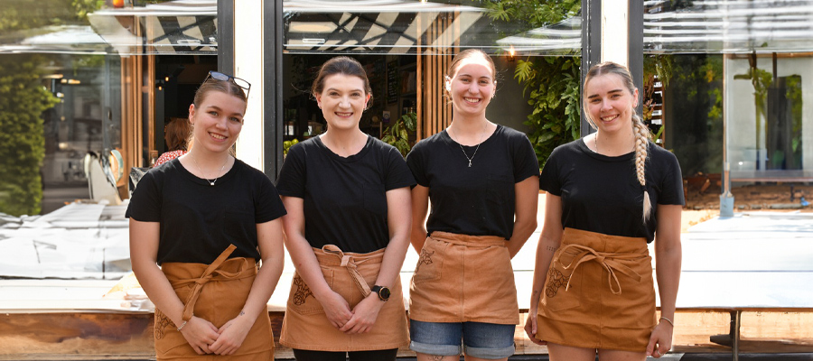 Four staff members of The Winey Cow standing outside the restaurant, smiling and wearing matching black shirts and brown aprons. The background shows the restaurant's outdoor seating area and entrance.