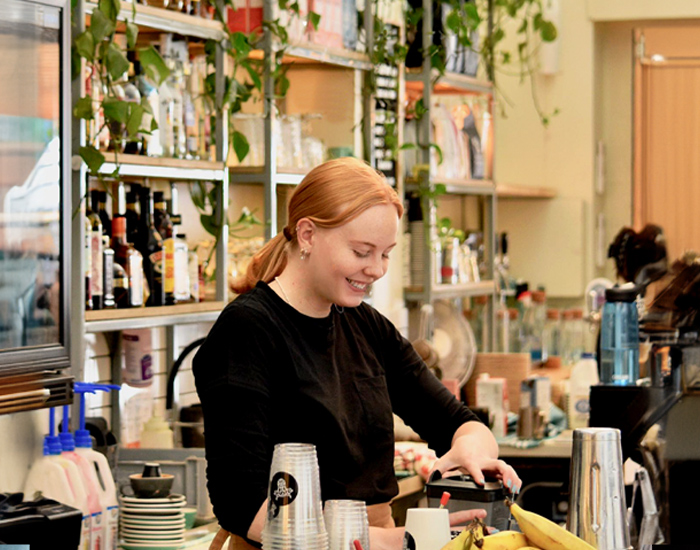 Smiling barista preparing a drink behind the counter at The Winey Cow, surrounded by shelves stocked with bottles, plants, and various barista tools. The environment appears busy and well-organized.