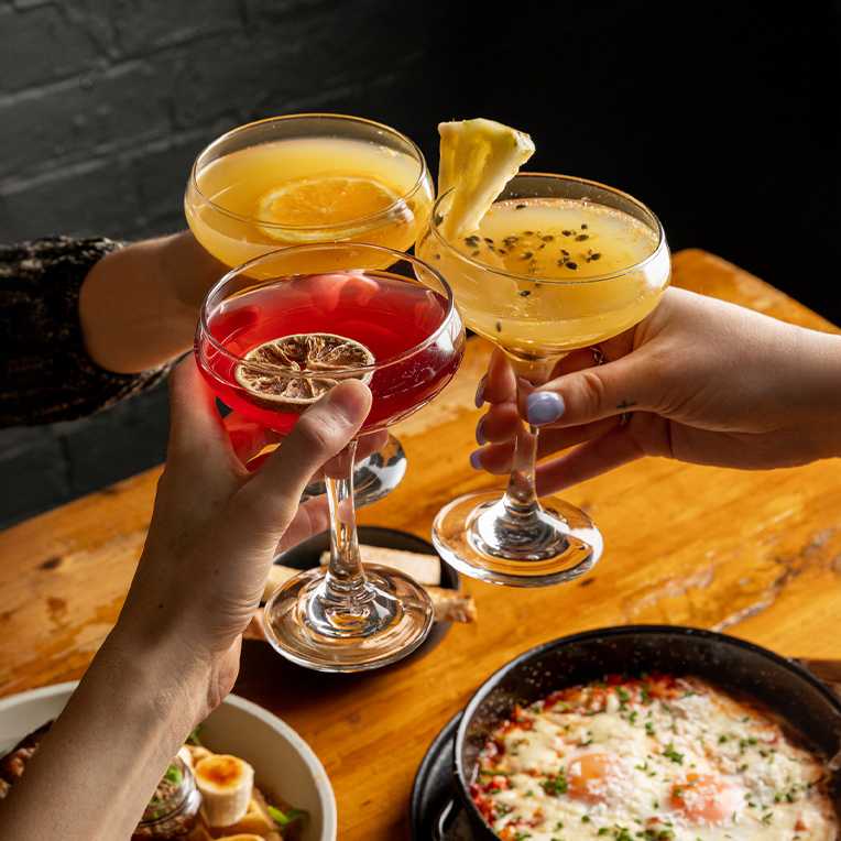 A vibrant image of 3 hands making a cheers with three different flavours of mimosas over a table of bunch food.