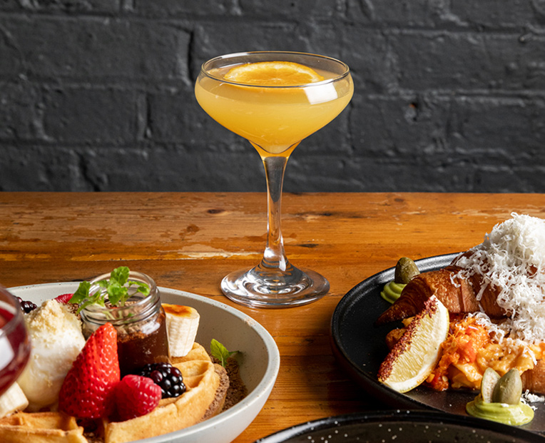 A single yellow mimosa on a wooden table next to two vibrant plates of breakfast dishes.