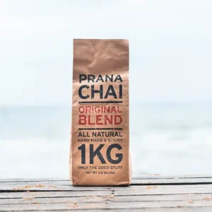 Bag of Prana Chai Original Blend, labeled as "All Natural, Handmade & Sticky, 1 KG," sitting on a wooden surface with a beach and ocean in the background. The packaging highlights its natural ingredients and artisanal quality.