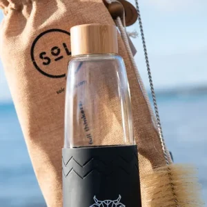 A glass water bottle with a bamboo top and a black silicone band around the bottom with The Winey Cow logo in white on the front paired with a carry case and scrub brush.