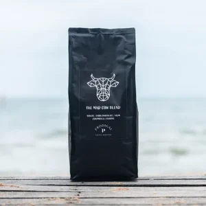 Bag of The Mad Cow Blend coffee by Prodigal Coffee Roasters, labeled with flavor notes of nougat, dark chocolate, and plum, sourced from Colombia and Ethiopia. The bag is black with a geometric cow logo, placed on a wooden surface with a beach and ocean in the background.