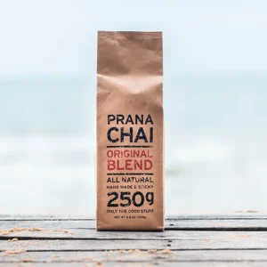 Bag of Prana Chai Original Blend, labeled as "All Natural, Handmade & Sticky, 250g," placed on a wooden surface with a beach and ocean in the background. The packaging emphasizes its natural ingredients and artisanal quality.