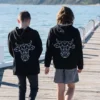 The back of two young boys walking on a dock wearing black sweatshirts that have The Winey Cow logo in white on the back.