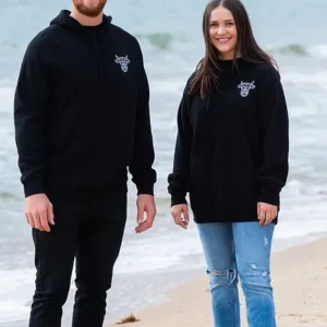 A smiling man and woman both standing on the beach wearing black sweatshirts with The Winey Cow logo in white on the chest.