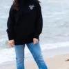 A young woman standing on the beach wearing a black sweatshirt with The Winey Cow logo in white on her chest.