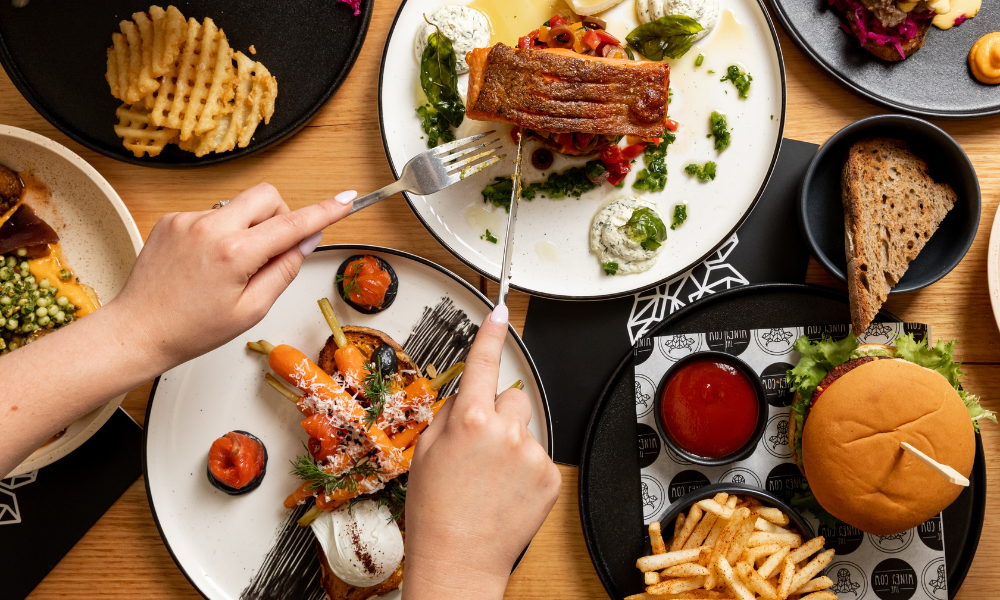 Bright birds-eye-view photo of a table full of different breakfast and lunch meals. Two hands are reaching in with a knife and folk to cut a piece of salmon.