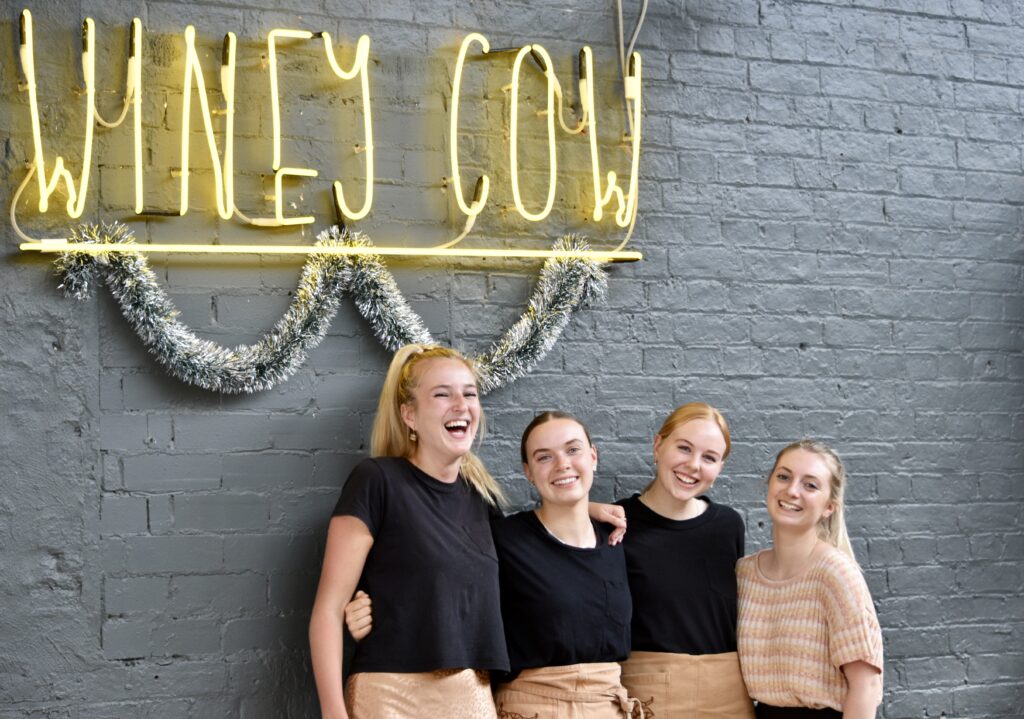 Four staff members at The Winey Cow smiling directly at the camera as they stand under a tinsel decorated 'Winey Cow' neon sign, ready for a Christmas Party.