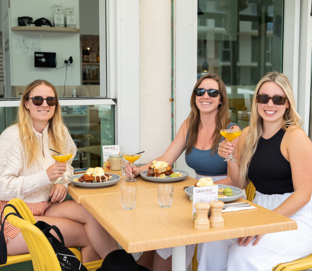 A bridge picture of three ladies sitting at a table with delicious brunch meals and mimosas in hand, enjoying a bottomless brunch experience.