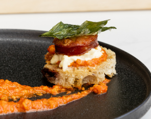 A canape of chorizo, Stracciatella cheese, romesco sauce, fried basil, and olive bread on a black plate.